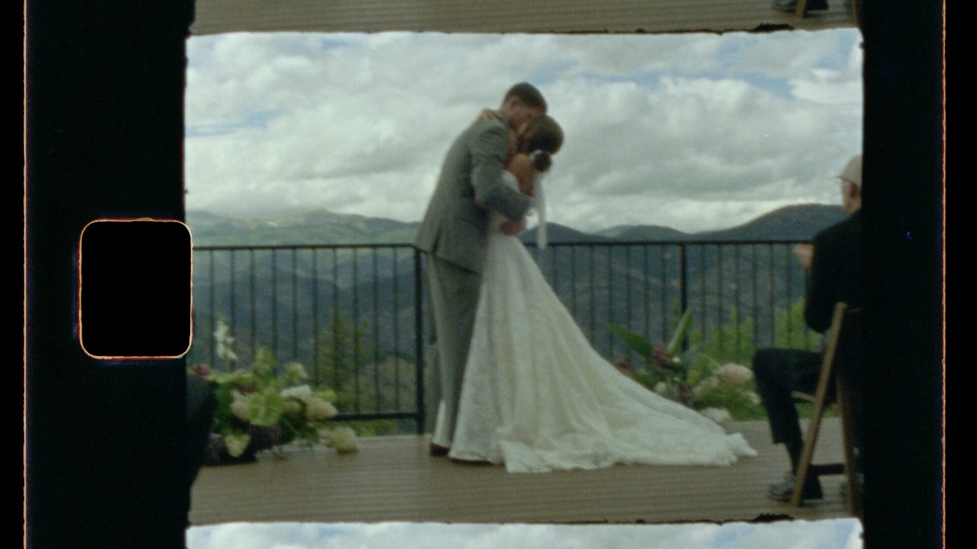 Super 8mm wedding film frame — ceremony first kiss on a mountain deck, Colorado peaks behind them, film frame border and organic grain visible. Shot on Kodak Super 8mm film stock.